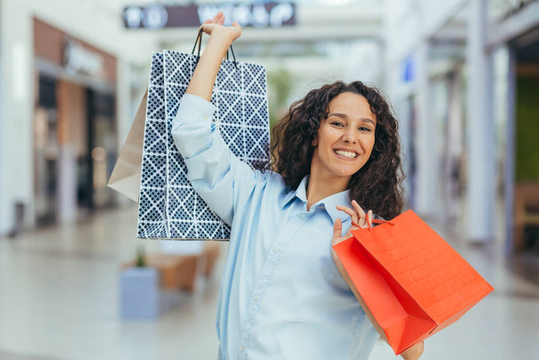 Ferienverkauf. Schwarzer Freitag. Glückliche junge hispanische Frau beim Einkaufen in einem Supermarkt. Sie hält bunte Papiereinkaufstüten in den Händen, hebt sie hoch, jubelt, lächelt in die Kamera.. - Foto, Bild