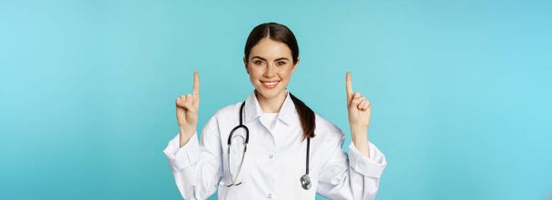 Πορτρέτο της αυτοπεποίθηση νεαρή γυναίκα γιατρός, ιατρός εργαζόμενος σε παλτό, δείχνοντας τα δάχτυλα πάνω και χαμογελώντας, δείχνει την ιατρική διαφήμιση, κλινική promo, τυρκουάζ φόντο. - Φωτογραφία, εικόνα