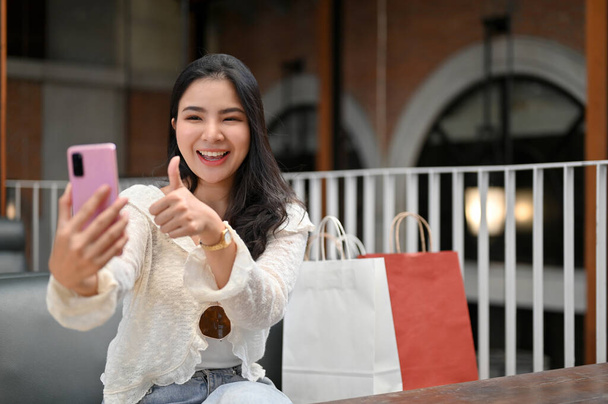 Overjoyed young Asian woman with her shopping bags showing thumb up, enjoy talking on a video call with her friend through her phone while relaxing at the cafe in the shopping mall. - 写真・画像