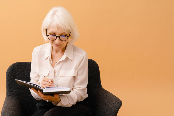Gyönyörű mosolygós idős nő üzleti öltönyben ül a karosszéken és jegyzeteket ír elszigetelt barna háttér - Fotó, kép