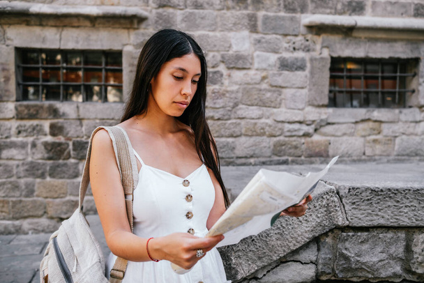 Donna latinoamericana in abito bianco con zaino esaminando mappa cartacea fuori vecchio edificio in pietra sulla strada di Barcellona, Spagna - Foto, immagini