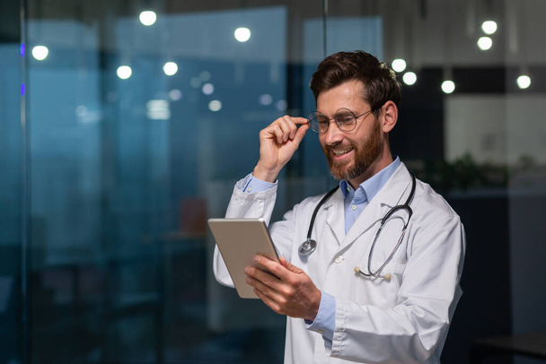 A young male doctor, intern, intern is standing in an office in a hospital in a white coat and with a stethoscope. He works on a tablet, holds his glasses in his hand, and smiles. - 写真・画像