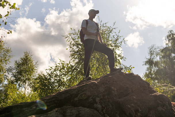 Giovane escursionista con zaino e bastoncini da trekking in piedi sulla scogliera e guardando le montagne in estate all'aperto - Foto, immagini
