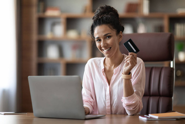 Attractive millennial brunette woman employee paying for goods and services on Internet while working at home office, sitting at desk in front of laptop, showing credit card and smiling, copy space - Foto, Imagem