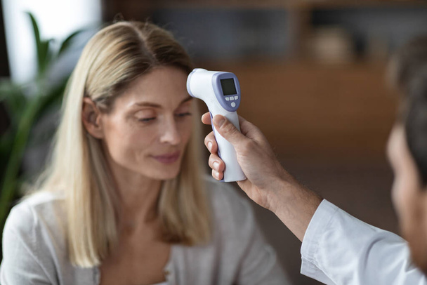 赤外線温度計を使用した女性患者のための認識できない医師の身体温度をチェックします。, covid-19流行中にクリニックでのチェックアップを持っている病気の中年女性,選択的フォーカス - 写真・画像