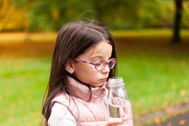Cute girl with long hair and glasses drinks water from a plastic bottle on a blurred autumn park background - Photo, Image