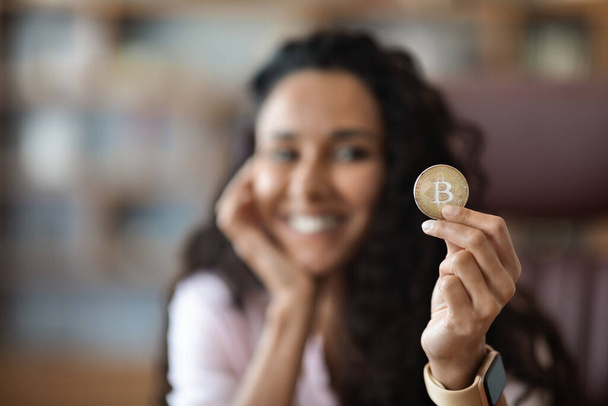 Gros plan portrait de jeune femme brune bouclée montrant Bitcoin et souriant, accent sélectif sur la pièce d'or, fond flou, intérieur de bureau, espace de copie, concept crypto-monnaie - Photo, image