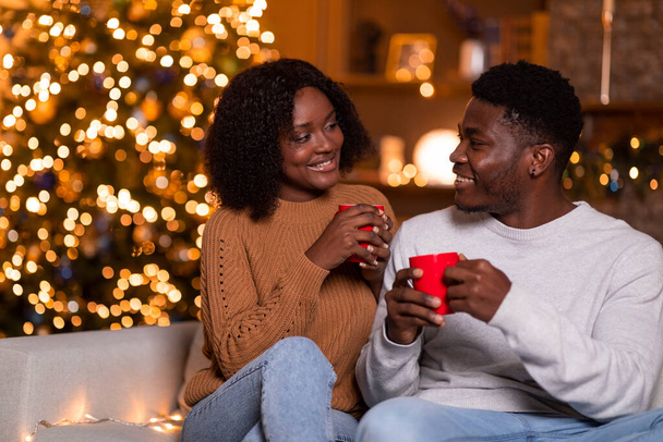 Smiling young african american guy and lady with cups of hot drink, enjoy holiday, free time, sit on sofa in living room interior with glowing garlands. Evening with coffee or cocoa at home together - Photo, Image