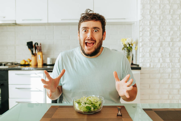 A young man is sitting in the kitchen with a bowl of salad and does not want to eat it, screaming in despair. A man on a diet eats vegetarian food. Only salad. Healthy eating concept. Stop diet. - Foto, immagini