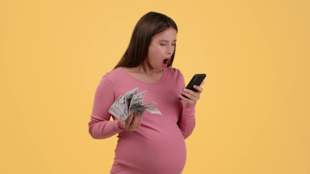 Ganar en línea para todos. Mujer embarazada joven mirando el teléfono inteligente, disfrutando de beneficios financieros, la celebración de ventilador de dinero, fondo de estudio naranja, cámara lenta, espacio libre - Metraje, vídeo