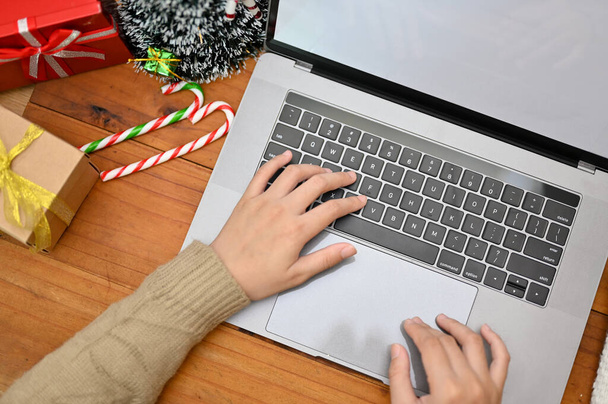 Nad głową, Kobieta w przytulnym swetrze wpisująca na klawiaturze laptopa, pracująca nad swoim projektem, korzystająca z notebooka na swoim miejscu pracy ze świątecznym wystrojem. - Zdjęcie, obraz