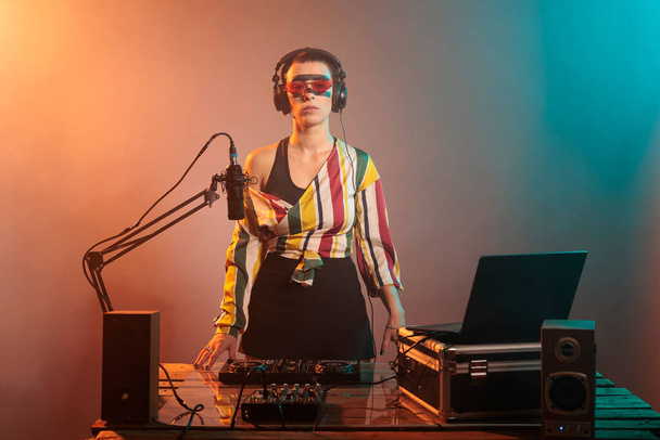 Lächelnder junger Musiker mit cooler Sonnenbrille auf einer Sutdio-Party, der Techno-Musik an Plattentellern mixt und Schallplatten benutzt. Künstler bei einem Auftritt in einem Nachtclub mit Audioanlage und Mischpult. - Foto, Bild
