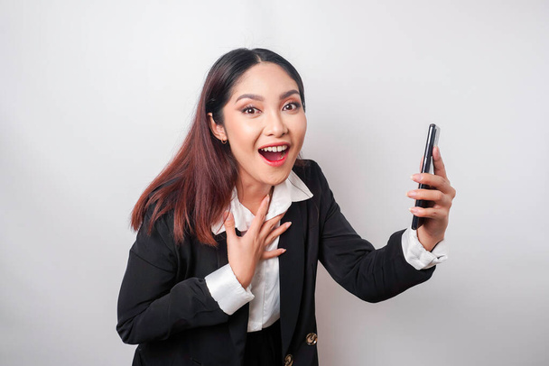 Surprised Asian businesswoman wearing black suit holding her smartphone, isolated by white background - Φωτογραφία, εικόνα