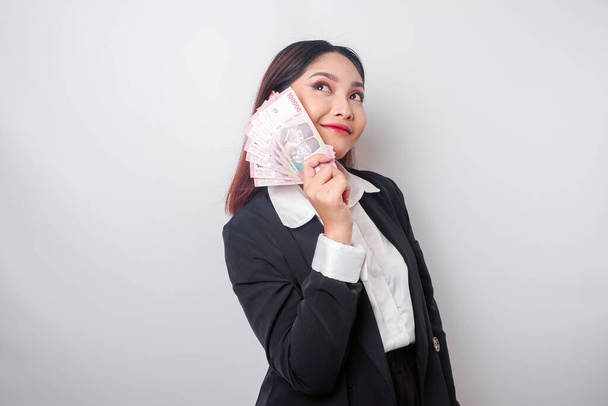 A thoughtful young woman is wearing black suit and holding cash money in Indonesian rupiah isolated by white background - 写真・画像