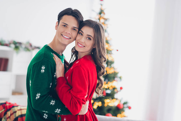 Photo of young couple together cuddles wear ugly sweaters enjoy spend time together christmastime near fir tree lights illumination indoors. - Fotoğraf, Görsel