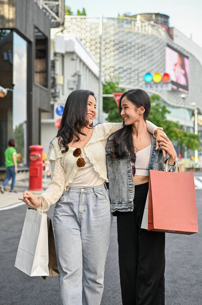 Δύο χαρούμενες και χαρούμενες νεαρές Ασιάτισσες που κουβαλάνε τις τσάντες τους, περπατάνε μαζί στον εμπορικό δρόμο, απολαμβάνουν τα ψώνια τους το Σαββατοκύριακο.. - Φωτογραφία, εικόνα