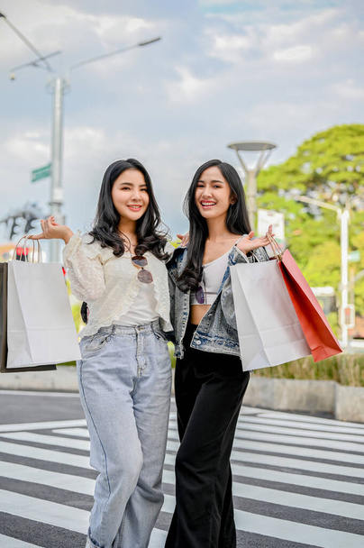 Ritratto, Due affascinanti e graziose giovani donne asiatiche che trasportano le loro borse della spesa, in piedi sul marciapiede, dopo aver goduto di un weekend di shopping insieme. - Foto, immagini