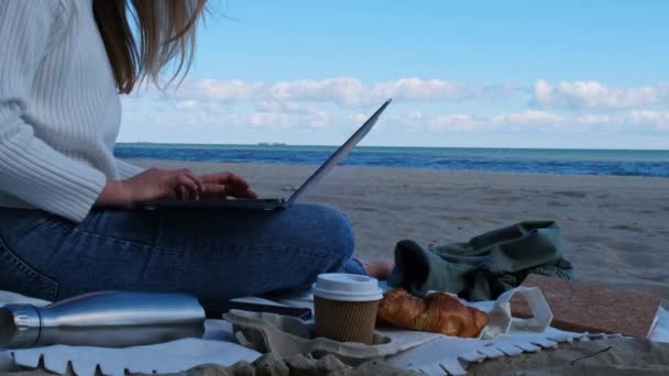 海の景色の隣のビーチでノートパソコンで働く若い女性。コーヒーを飲み、クロワッサンを食べる。学生勉強は暖かい秋の海岸の屋外で動作します。フリーランス  - 映像、動画