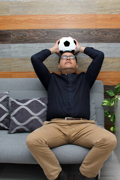 眼鏡をかけたラテン系の大人の男は彼のボールと彼のソファの上でテレビでサッカーゲームを見て、怒って、悲しいサッカーゲームを見て興奮します - 写真・画像