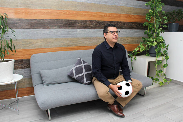 Latino adult man with glasses watches soccer game on TV on his sofa with his ball and gets excited, angry, sad watching the football game - Photo, Image