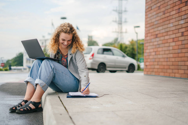 Διεθνής φοιτητής που σπουδάζει online, κρατώντας σημειώσεις ενώ ακολουθεί τον καθηγητή σε βιντεοκλήση, κάθεται σε εξωτερικούς χώρους. Έννοια της εκπαίδευσης, πανεπιστημιούπολη, έφηβος. Άνθρωποι - Φωτογραφία, εικόνα