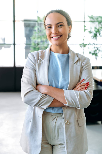 Κάθετη προσωπογραφία μιας όμορφης κομψής νεαρής γυναίκας μικτής φυλής, επιτυχημένης εργαζόμενης εταιρείας, ceo, στέκεται στο γραφείο της με σταυρωμένα χέρια, κοιτάζοντας την κάμερα, χαμογελώντας φιλικά - Φωτογραφία, εικόνα