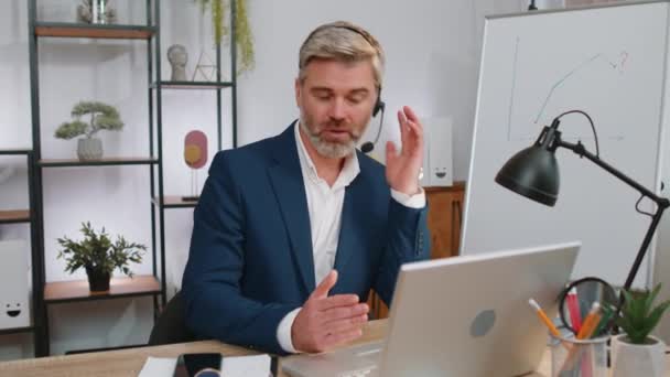 Mature business man working on laptop wearing headset, freelance worker call center or support service operator helpline, having pleasant talk with client or colleague communication support at office - Footage, Video
