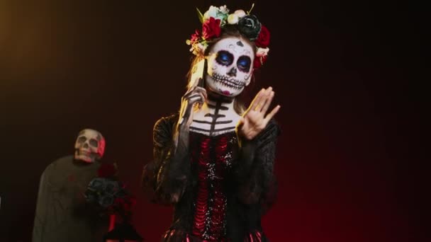 Spooky goddess talking on phone call with smartphone, wearing flowers crown and cavalera catrina halloween costume. Woman dressed as santa muerte to celebrate mexican culture holiday. Handheld shot. - Footage, Video
