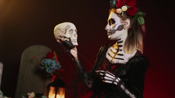 Модель Санта-муэрте флиртует с черепом и чёрными розами, носит традиционное искусство тела, чтобы отпраздновать мексиканский праздник. Выглядишь как богиня смерти на священном ритуале. Портативный выстрел. - Кадры, видео
