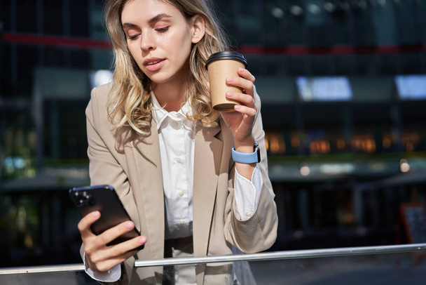 Stijlvolle corporate vrouw in pak, drinkt haar koffie afhaalmaaltijden en maakt gebruik van mobiele telefoon app. - Foto, afbeelding