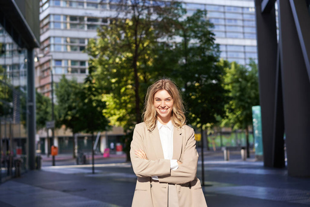 Portrait de jeune femme d'affaires confiante en costume beige, avocat d'entreprise ou vendeuse debout à l'extérieur de l'immeuble de bureaux dans la rue et souriant. - Photo, image