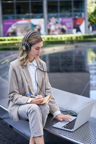 Portrait of businesswoman being on a meeting online outside office, listening in headphones, looking at laptop, working outdoors. - Photo, Image
