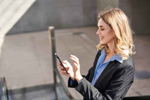 Ritratto di giovane donna d'affari in scala mobile, con smartphone e sorriso, toccare il telefono cellulare, indossare tuta. - Foto, immagini