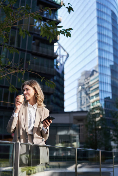 Femme d'affaires enthousiaste boit son café à emporter dans la rue, tient son téléphone portable, debout en costume beige, tenue de bureau. - Photo, image