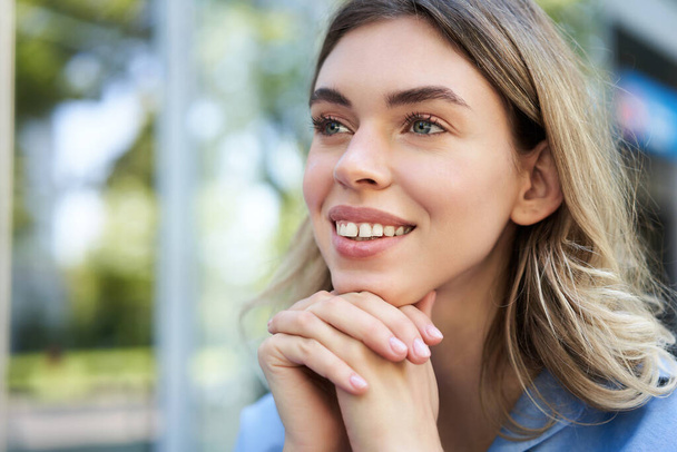 Close up portrait of young woman, looking hopeful and smiling, lean head on hands, thinking, sitting outdoors. - Foto, Bild