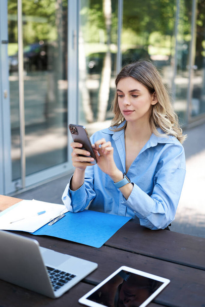 Portrait of businesswoman working outside in park, sitting with laptop, work documents and using mobile phone, text message, reading on smartphone. - Фото, зображення