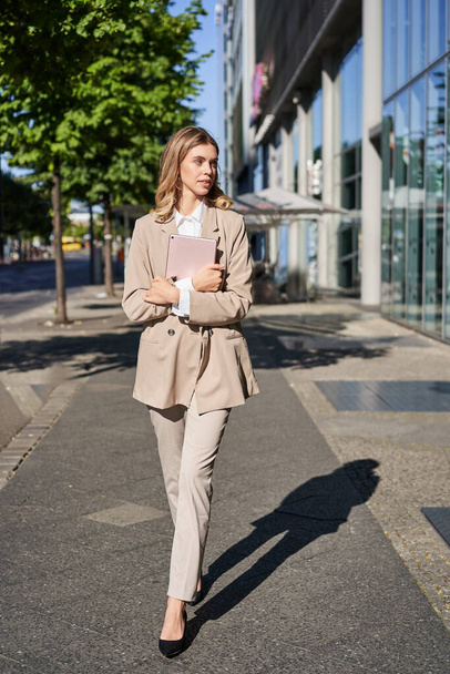 Vertikale Aufnahme einer Geschäftsfrau, die mit digitalem Tablet auf der Straße läuft, zur Arbeit geht und beigen Anzug und High Heels trägt. - Foto, Bild