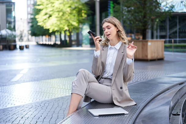 İş kadını şehir merkezinde oturuyor ve cep telefonuyla çalışıyor. Dizüstü bilgisayarı yanında ve iş kıyafetleri giyiyor.. - Fotoğraf, Görsel
