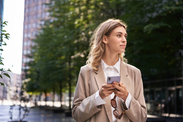 Portret van zakenvrouw in pak, houdt haar mobiele telefoon tijdens het lopen op straat. - Foto, afbeelding
