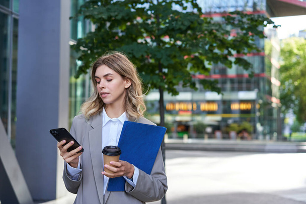 Businesswoman drinking coffee and holding documents with smartphone, going to work in office, standing outdoors in city center. - Photo, Image