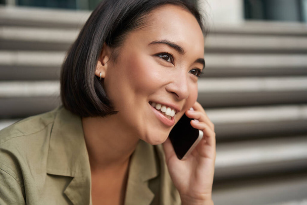 Communication. Jeune femme asiatique parlant sur un téléphone mobile, ayant un appel téléphonique, en utilisant un smartphone tout en étant assis sur la rue. - Photo, image