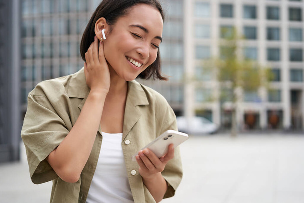 Χαμογελαστή Ασιάτισσα ακούει μουσική με ασύρματα ακουστικά, κοιτάζει το τηλέφωνό της, διαλέγει μουσική ή podcast. Νεαρή γυναίκα καλεί κάποιον, με ακουστικά. - Φωτογραφία, εικόνα