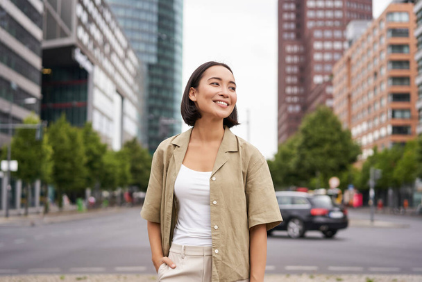 Szabadtéri felvétel fiatal, mosolygós ázsiai nőről, aki nappal az utcán áll, körülnéz, boldognak tetteti magát.. - Fotó, kép