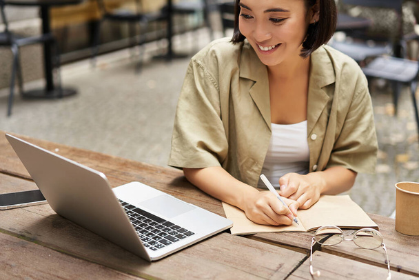 Retrato de una mujer joven que estudia en línea, sentado con el ordenador portátil, anotando, tomando notas y mirando la pantalla de la computadora, sentado en la cafetería al aire libre. - Foto, imagen