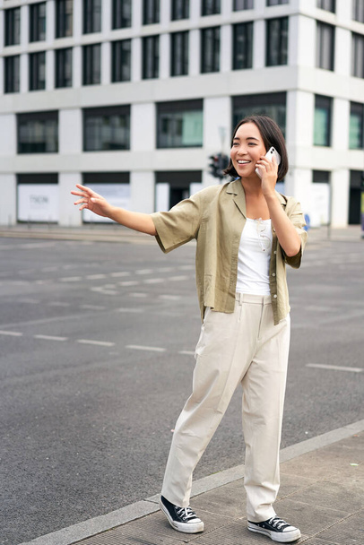 Young woman waving to taxi driver, calling or talking on mobile phone, order car ride, standing outdoors on street. - Foto, afbeelding
