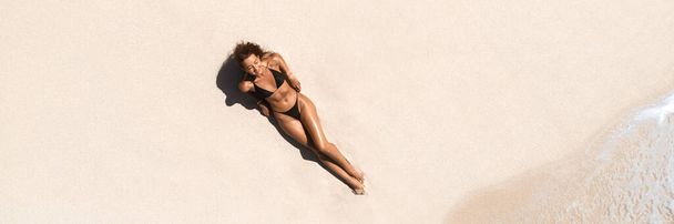Top antenne drone uitzicht op de vrouw in badpak ontspannen en zonnebaden op het strand wit zand in de buurt van de oceaan. Aantrekkelijk brunette meisje in zwarte bikini liggend aan een zandkust met palmblad. Prachtig gebruind - Foto, afbeelding