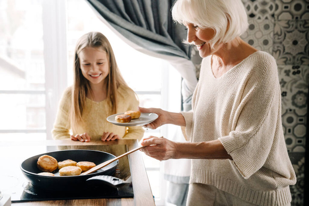 Happy grandmother and granddaughter cooking cheese pancakes together in kitchen at home - 写真・画像
