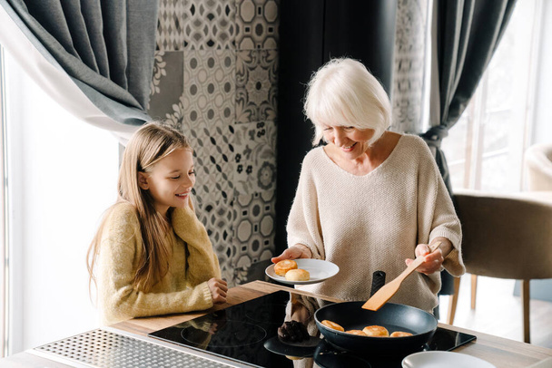 Happy grandmother and granddaughter cooking cheese pancakes together in kitchen at home - Фото, зображення