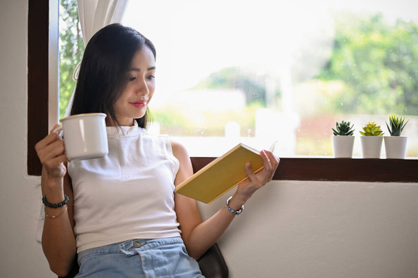 Mooie jonge Aziatische vrouw ontspannen thuis, zitten in de buurt van het raam, genieten met haar middag koffie en lees haar favoriete boek. - Foto, afbeelding