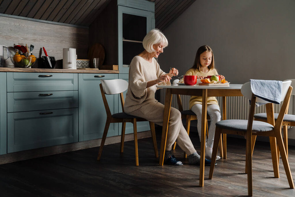 Happy senior woman having breakfast with her granddaughter in kitchen at home - Фото, зображення